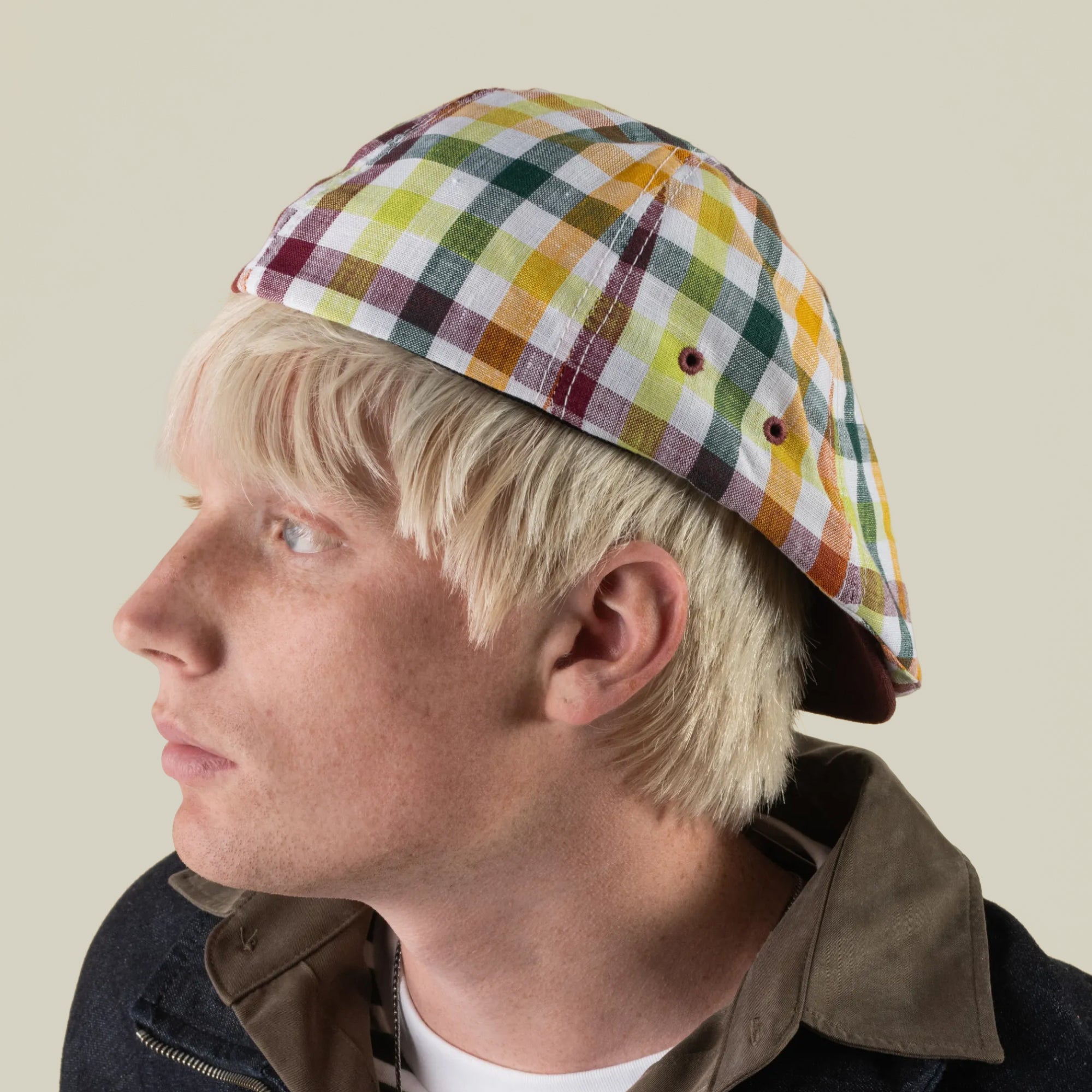 A person with light blond hair, wearing the Goorin Bros. Point Lobos multicolored plaid 6 Panel Ivy cap handmade in the USA and a dark jacket, looks to the left against a plain background.