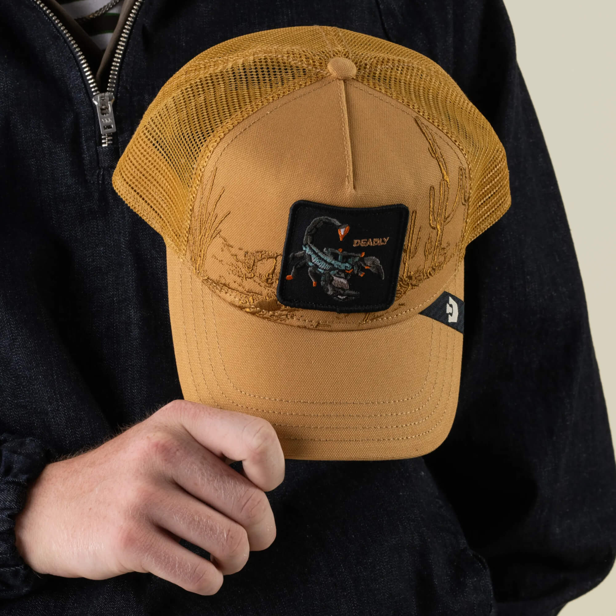 A person in a dark jacket holds the Badlands Scorpion trucker hat by Goorin Bros.—a tan mesh back cap featuring a scorpion patch labeled “DEADLY” with detailed desert illustrations.