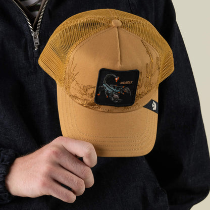A person in a dark jacket holds the Badlands Scorpion trucker hat by Goorin Bros.—a tan mesh back cap featuring a scorpion patch labeled “DEADLY” with detailed desert illustrations.