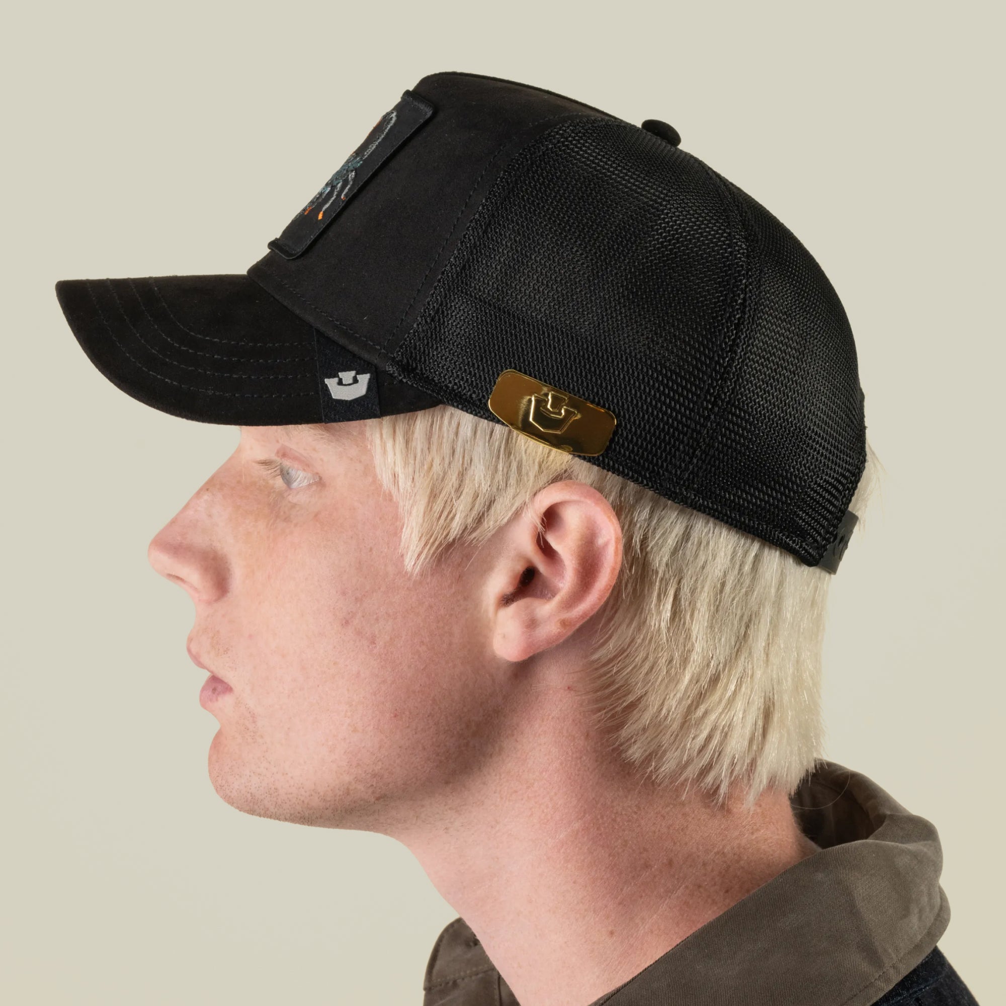 A person with light blond hair is shown in profile against a plain background, wearing a black mesh Goorin Bros. baseball cap called the Castle CapLoc, which features a gold-toned zinc alloy brim clip accessory on the side.