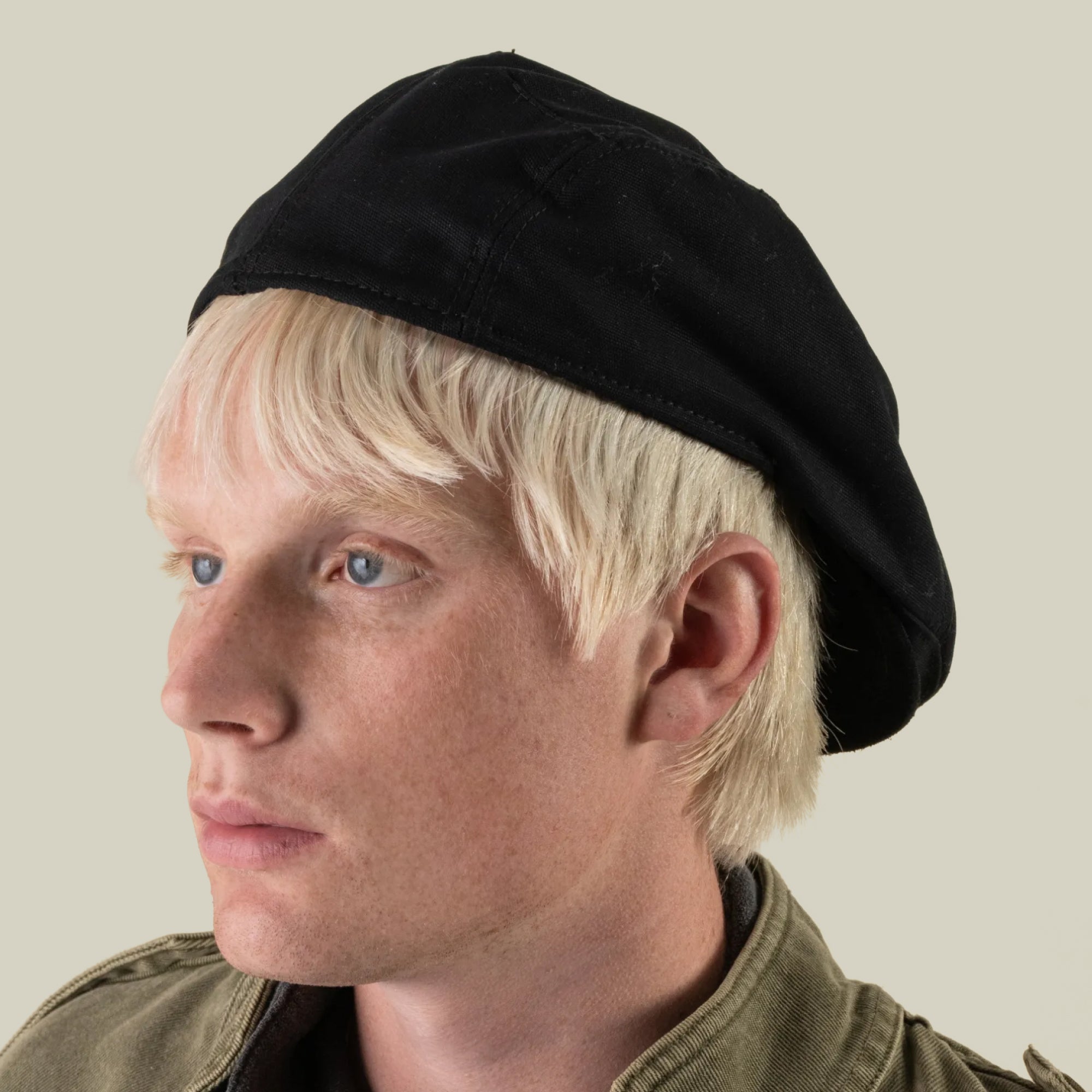 A person with light blond hair wearing the Goorin Bros. Heatherwood black cotton flatcap, made in the USA, and an olive green jacket, looks to the left against a plain light background.