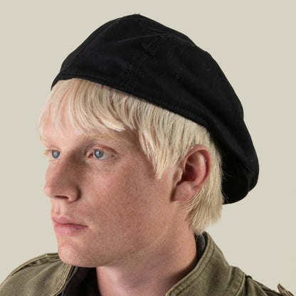 A person with light blond hair wearing the Goorin Bros. Heatherwood black cotton flatcap, made in the USA, and an olive green jacket, looks to the left against a plain light background.