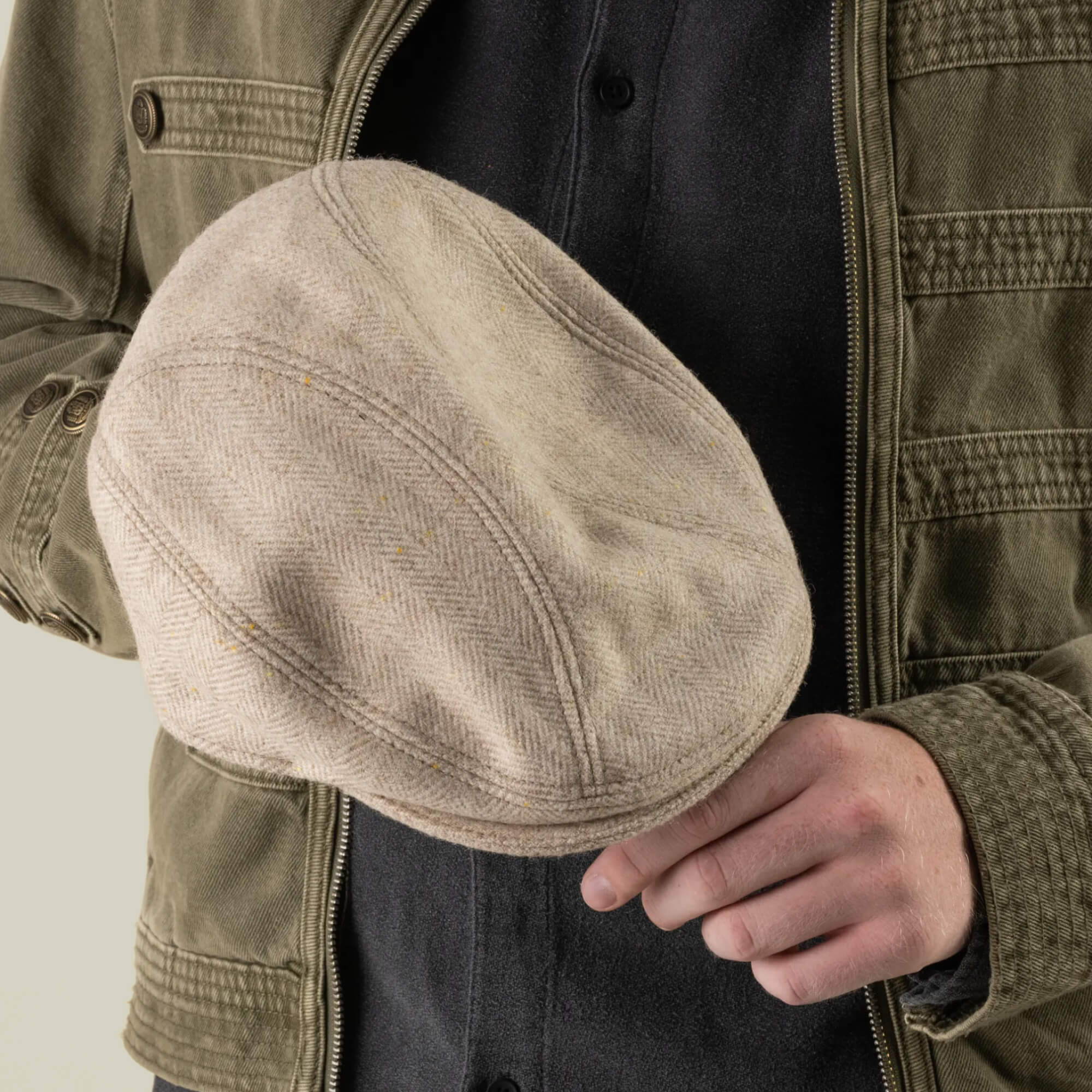 A person wearing an olive jacket and dark shirt holds the Goorin Bros. Iris Fields flat cap—hand-cut in the USA from 100% wool in a chalk cream color—in their right hand against a plain background.