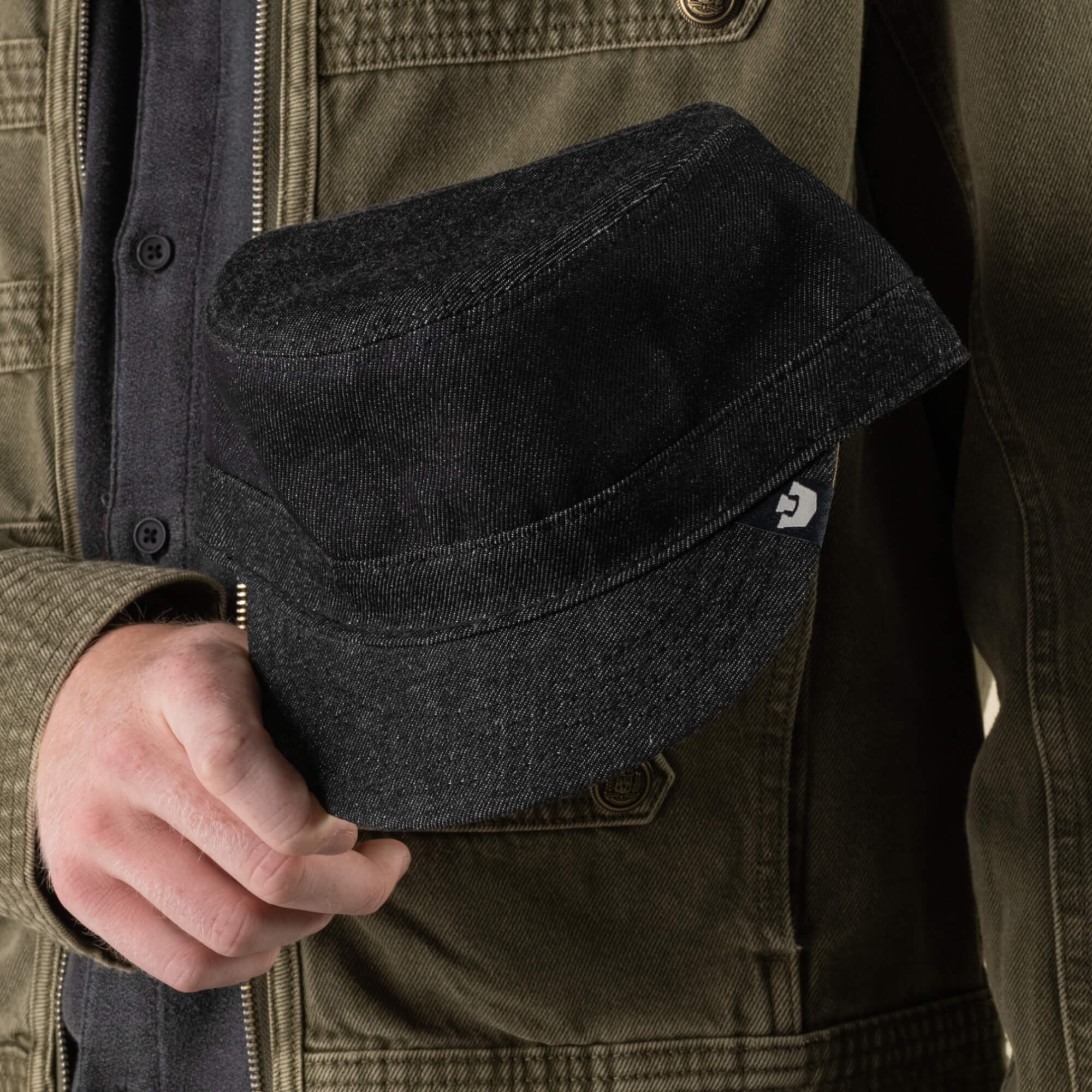 A person wearing an olive jacket holds the “Devonshire” black denim cotton cadet cap by Goorin Bros., hand-sewn in the USA, with a small visible logo tag on the side.