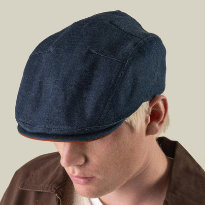 A person wearing the Goorin Bros. Ashfield, a dark blue hand sewn USA flat cap, along with a brown jacket, looking down against a plain light background.