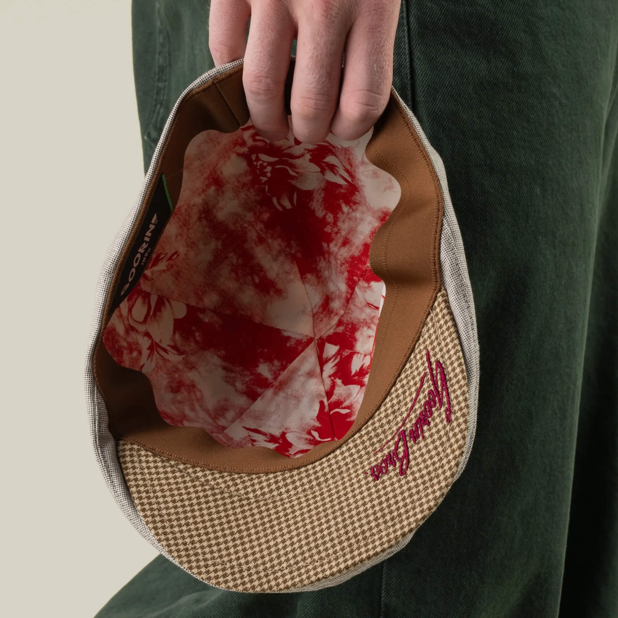 A person holds an open Goorin Bros. Mile Rock 6 Panel Ivy cap in beige houndstooth, revealing a red and white floral-patterned lining, the brand logo on the brim, and showcasing its hand cut and sewn craftsmanship.