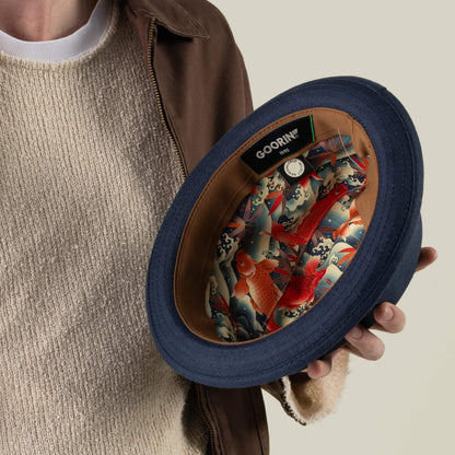 A person in a brown jacket and beige sweater holds the Charlie Pants by Goorin Bros., revealing its colorful fish-patterned lining and a tag labeled GOORIN, highlighting that the piece was hand-cut in the USA.