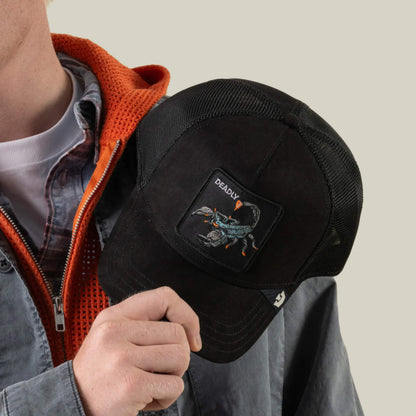 A person holds the Goorin Bros. Suede Scorpion, a black trucker hat featuring a suede front, breathable mesh back, and a patch on the front with an eagle and the word “DEADLY.” They’re dressed in layered shirts and a gray jacket with an orange hood.
