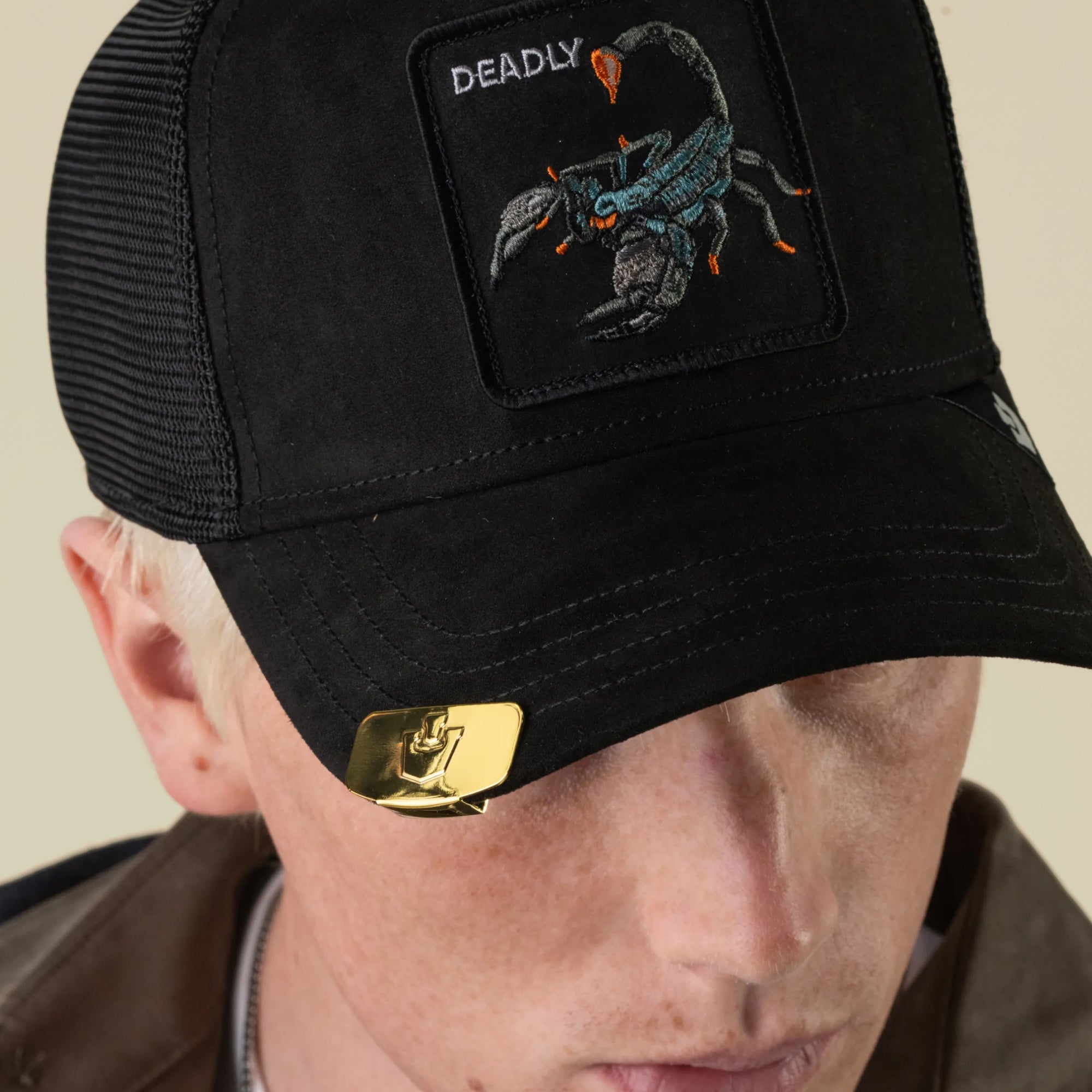 A person is wearing a black Goorin Bros. Castle CapLoc cap featuring a scorpion patch labeled DEADLY, with a gold zinc alloy Brim Clip accessory attached to the brim. The person is looking down, and their face is partially visible.