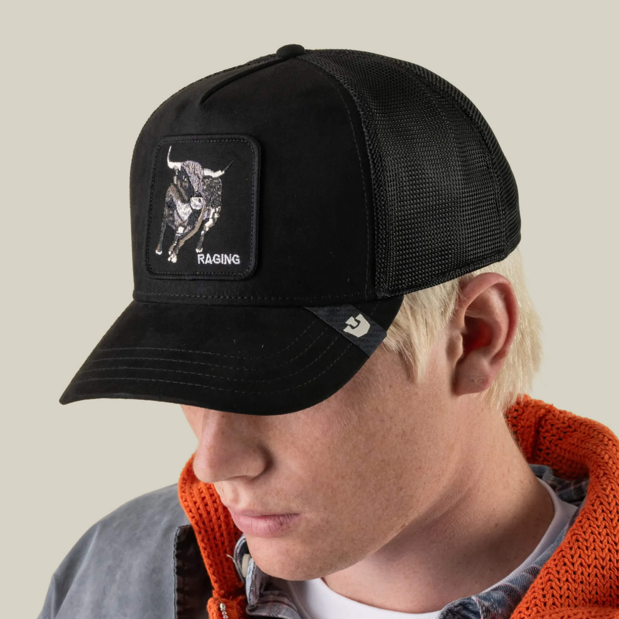 A person wearing a black mesh Goorin Bros. Suede Bull baseball cap featuring a bold RAGING Bull patch on the front and the word “RAGING,” paired with a grey jacket and an orange sweater, showcasing a striking approach to athletic-inspired fashion.