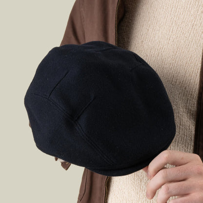 A person in a beige sweater and brown jacket holds the Goorin Bros. Cornerstone black flat cap, handmade in New Jersey, in their right hand against a plain background.