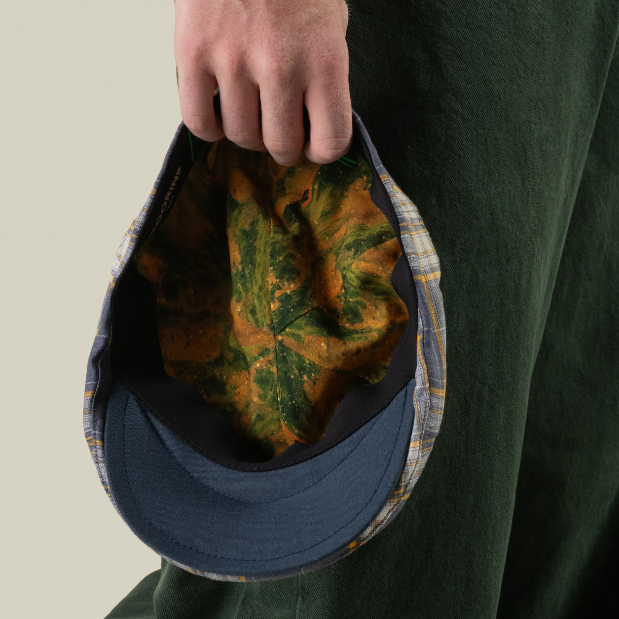 A person in green pants holds the Goorin Bros. Point Lobos cap, a handmade in USA 6-panel Ivy plaid hat featuring a blue brim and a leaf-patterned interior, with the cap facing downward.