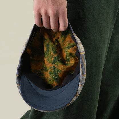 A person in green pants holds the Goorin Bros. Point Lobos cap, a handmade in USA 6-panel Ivy plaid hat featuring a blue brim and a leaf-patterned interior, with the cap facing downward.