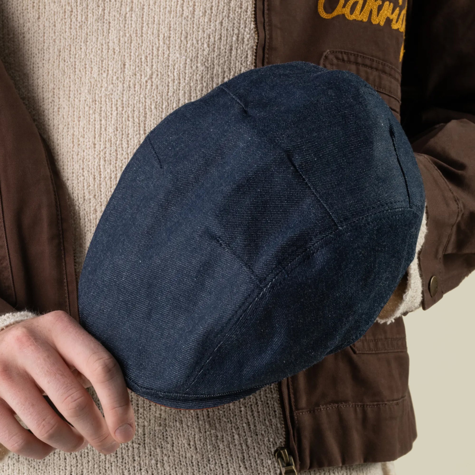 A person wearing a brown jacket and beige sweater holds the hand sewn Ashfield USA blue denim flat cap by Goorin Bros. in their left hand against a plain background.