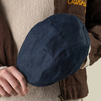 A person wearing a brown jacket and beige sweater holds the hand sewn Ashfield USA blue denim flat cap by Goorin Bros. in their left hand against a plain background.