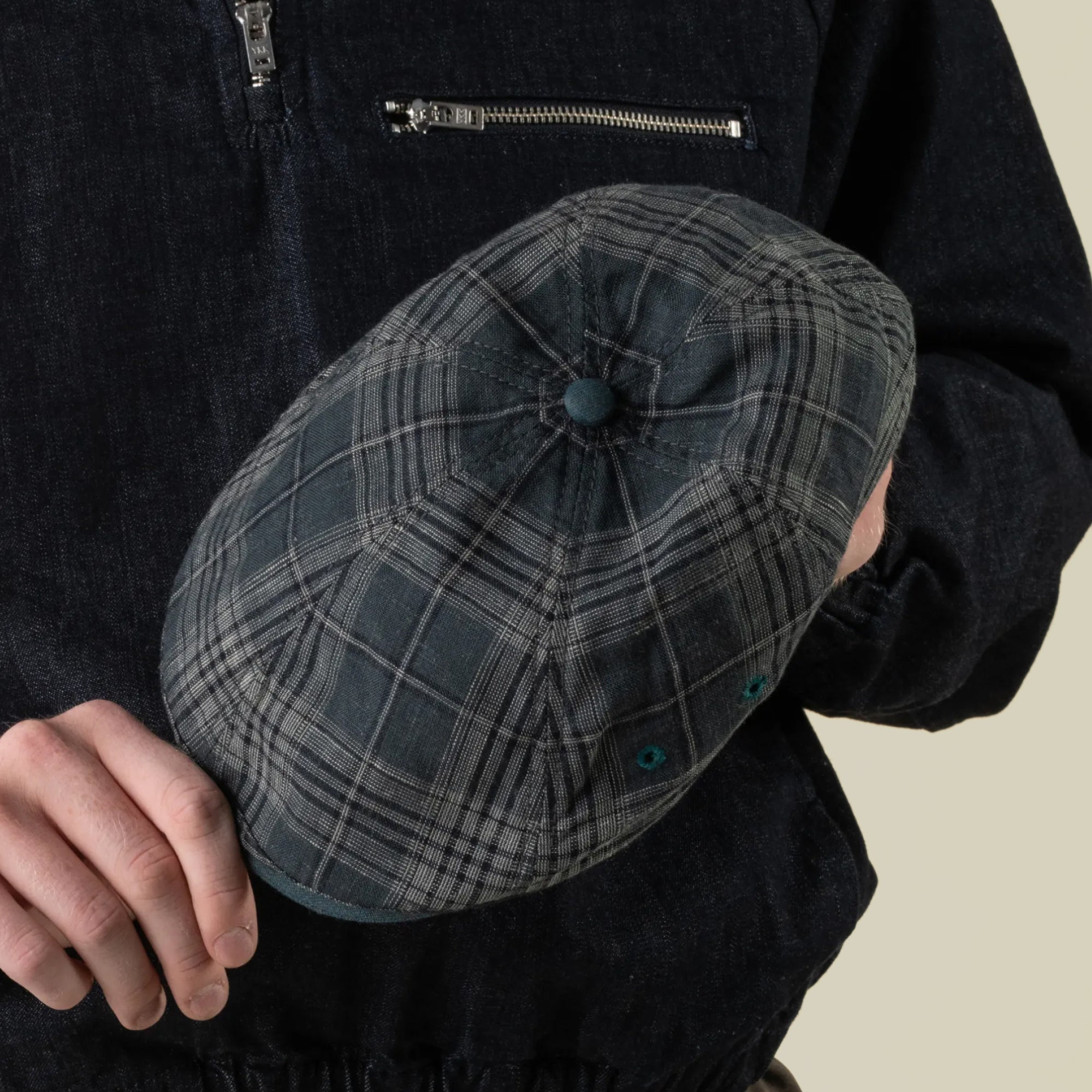 A person in a dark zippered jacket holds the Point Lobos 6 Panel Ivy flat cap by Goorin Bros.—handmade in the USA from a breathable cotton blend in grey plaid—with one hand against a plain background.