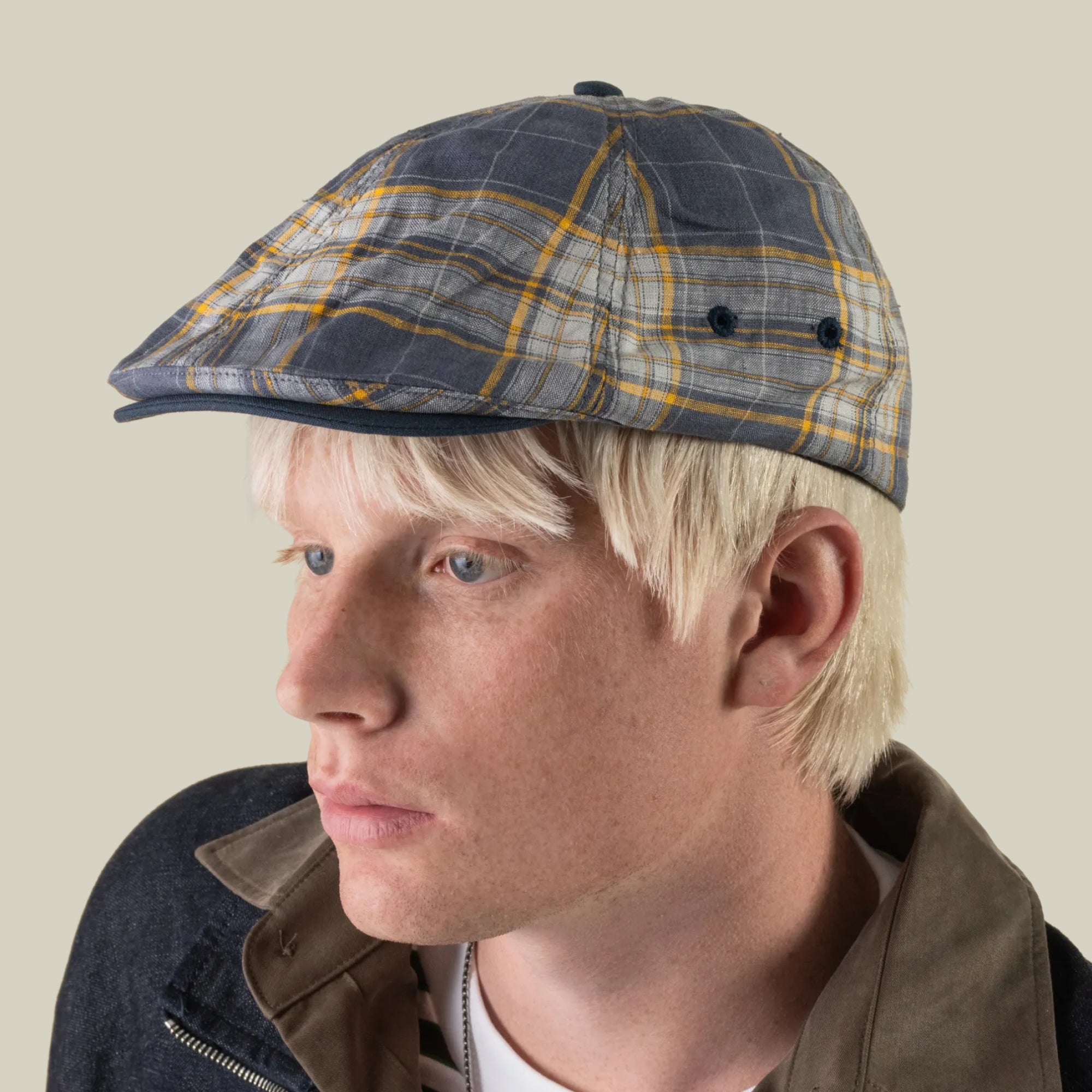 A person with platinum blonde hair wears the Goorin Bros. Point Lobos 6 Panel Ivy flat cap in gray and yellow plaid, handmade in the USA from a breathable cotton blend, paired with a dark jacket, looking to the left against a plain background.