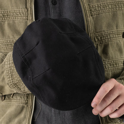 A person wearing a green jacket holds the Heatherwood flatcap by Goorin Bros., a black cotton cap with visible stitching, in front of their chest.