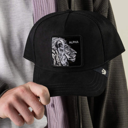 A person holds the Goorin Bros. Black Out Lion trucker hat, which features a black mesh design and a patch with an embroidered lion and the word “ALPHA” above it.