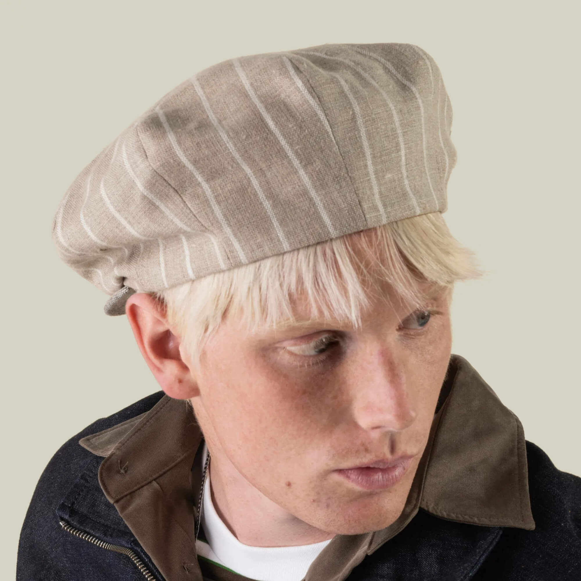 A person with blonde hair wears the Rincon Park beret by Goorin Bros.—a beige, pinstriped style hand cut and sewn from a linen blend—paired with a dark jacket, looking down and to the side against a plain background for a Gatsby-inspired silhouette.