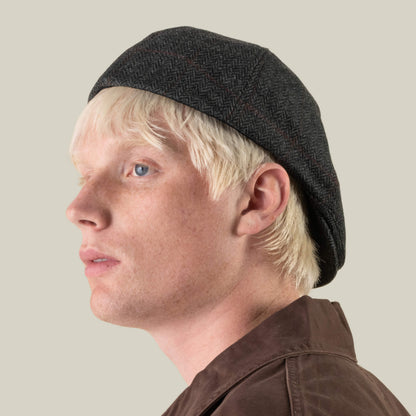 A person with pale skin and blonde hair wears the Goorin Bros. “Harry Bones” dark, textured 100% wool beret and a brown jacket, looking to the left against a neutral background.