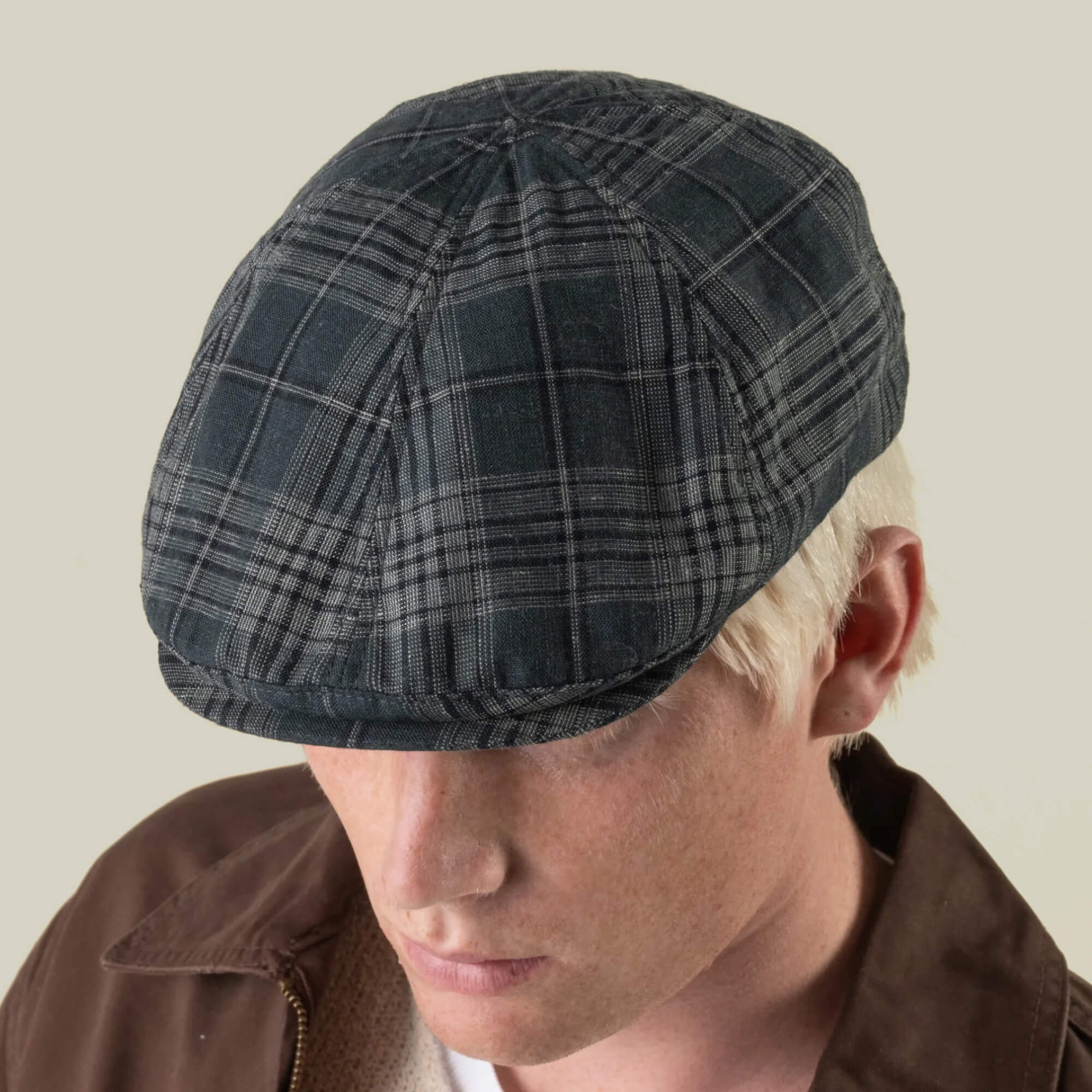 A person with light skin and blond hair wears the Goorin Bros. Glenwood Grove—a dark plaid flat cap, hand-cut in the USA with a premium headwear chassis—along with a brown jacket, looking downward against a plain background.