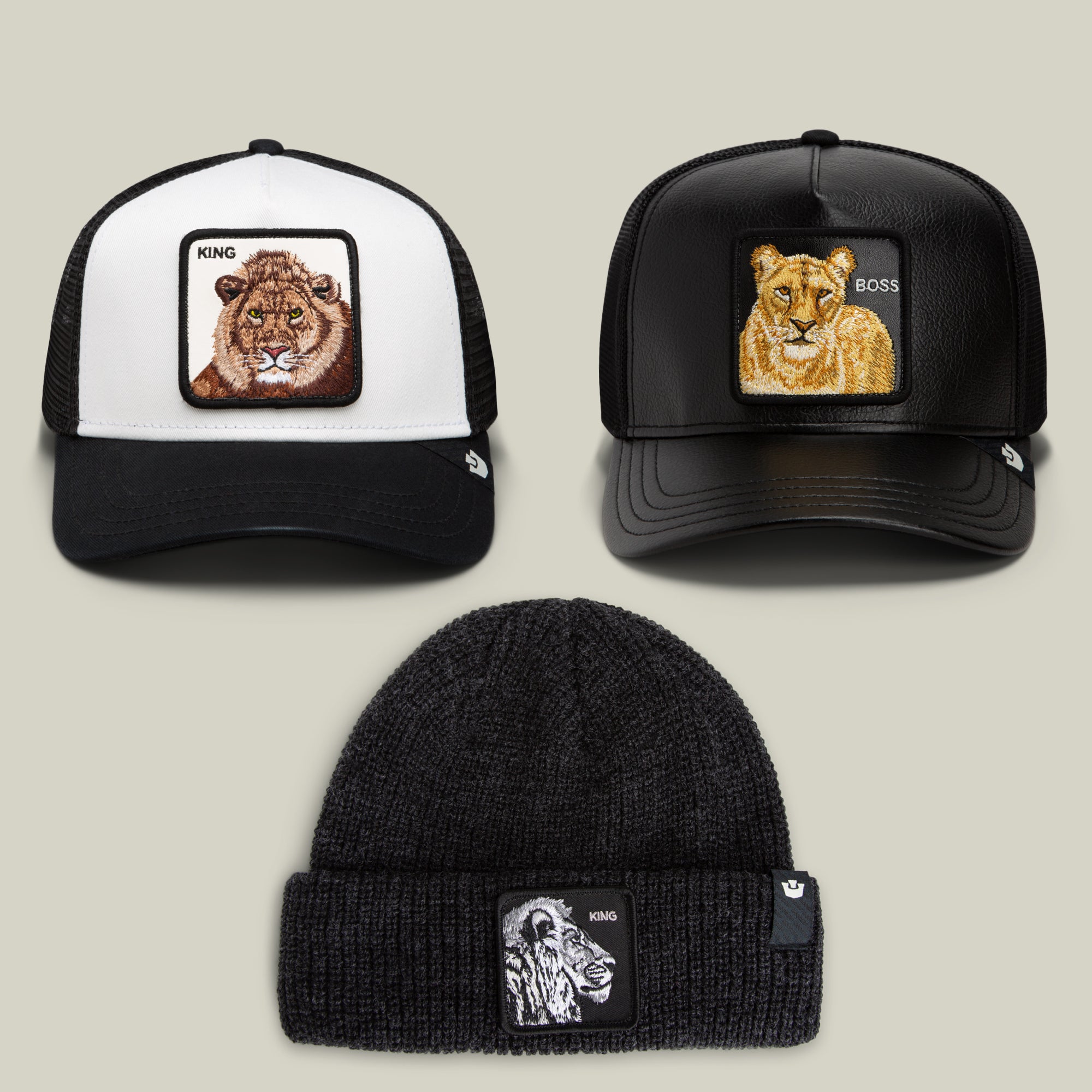 Three standout pieces from the Bend the Knee Bundle by Goorin Bros.—a lion trucker hat with a bold King patch, a lioness trucker hat featuring a Boss patch, and a black knit beanie adorned with a gorilla King patch—are artfully arranged on a neutral background.