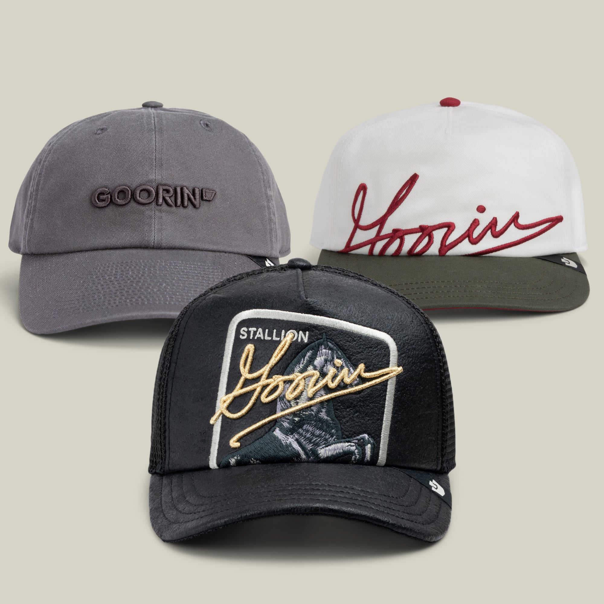 Here’s a rewritten sentence using the given product data: The Brandwear Bundle by Goorin Bros. includes three hats: a grey dad cap, a white and green cap with maroon cursive script, and a black mesh trucker hat featuring a Stallion patch with gold script.