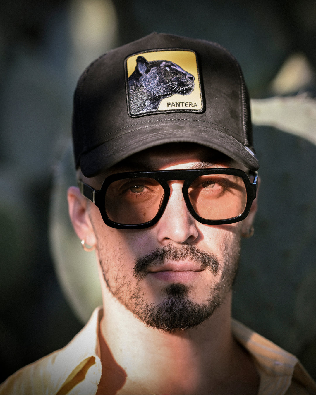 Man with a dark beard and mustache wearing large brown sunglasses and a black Goorin trucker hat featuring a panther patch that reads Pantera.