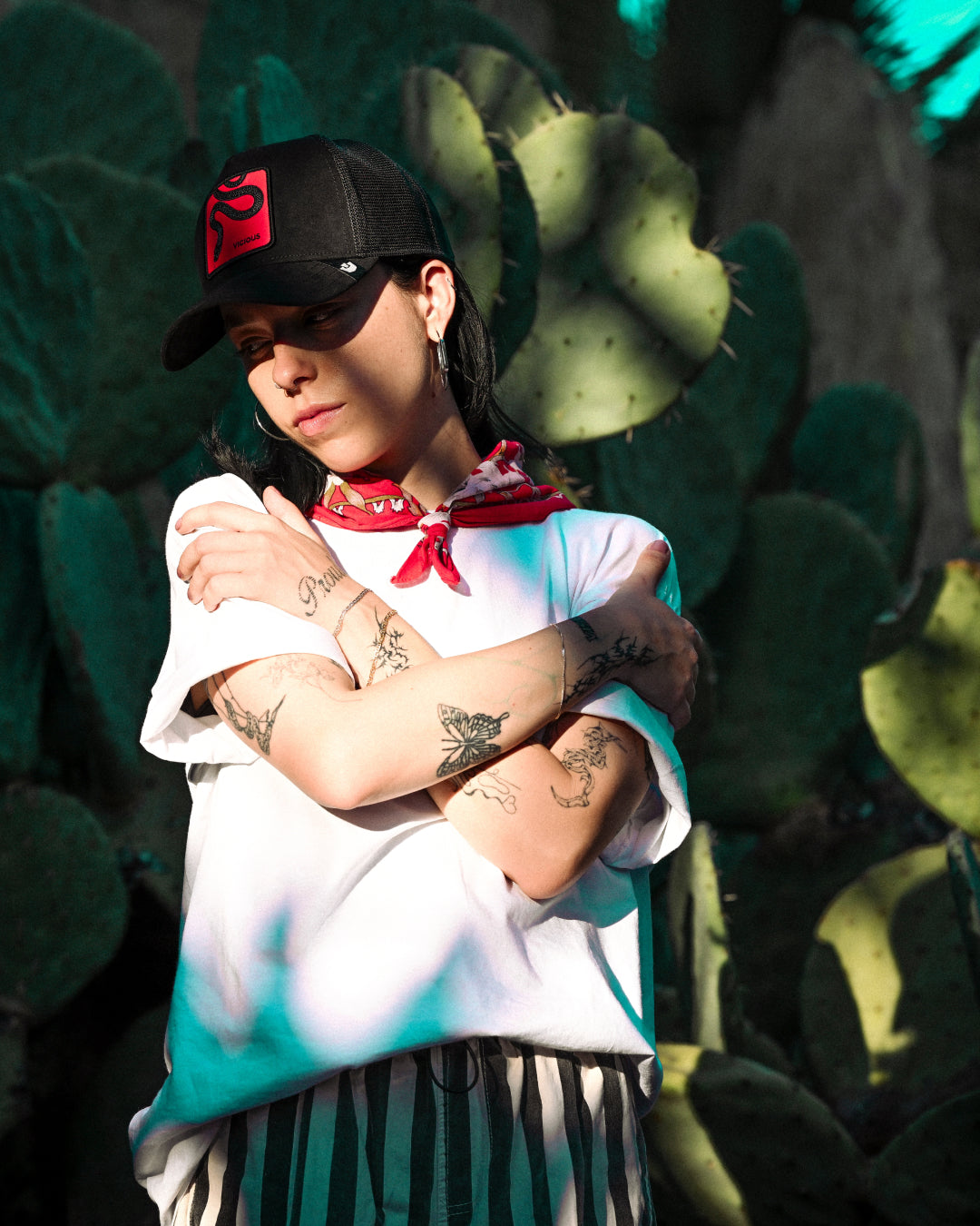 A person wearing a Goorin black cap, white shirt, and striped pants stands in front of cactus plants, arms crossed and eyes closed, with tattoos visible on their arms.