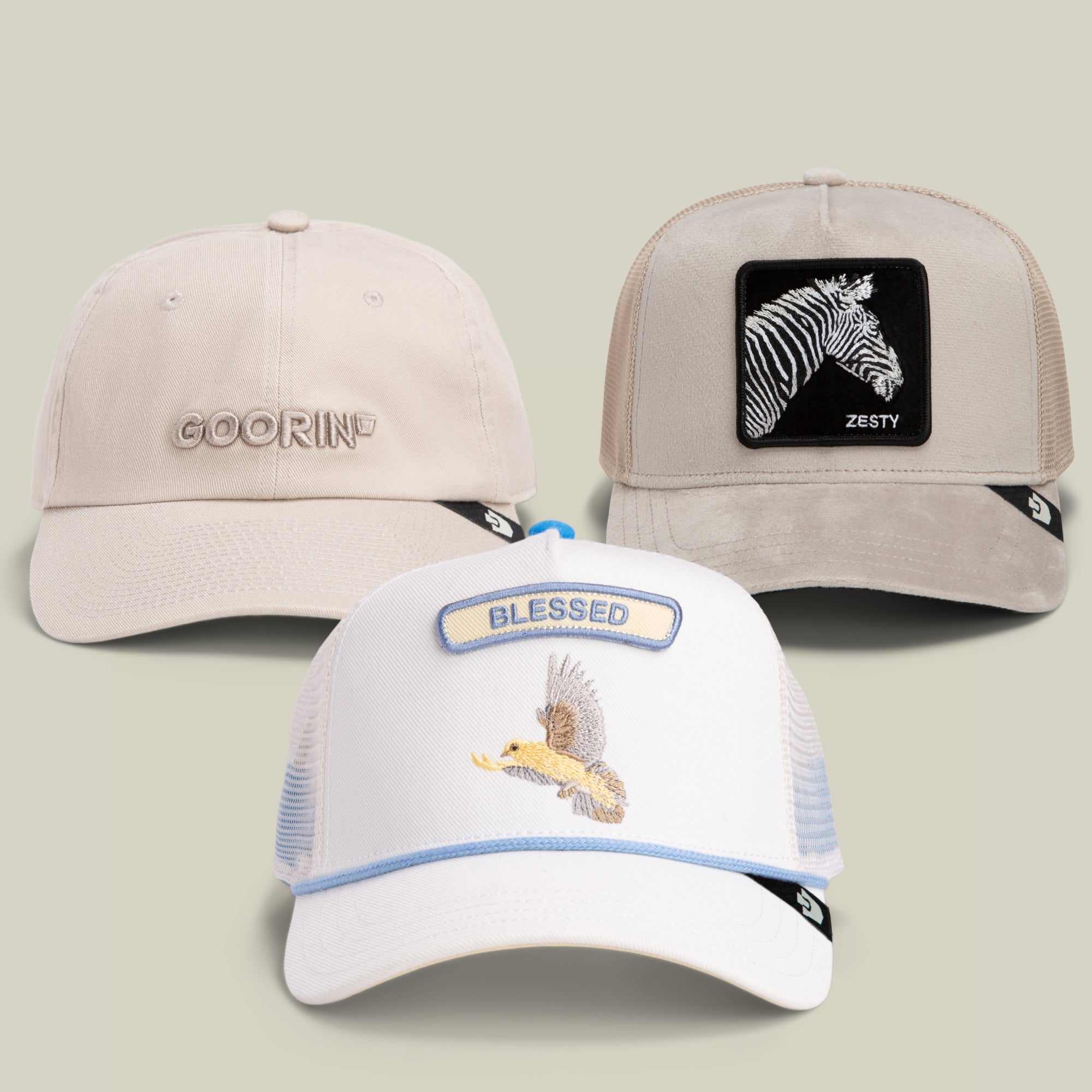 The Mr. Brightside Bundle by Goorin Bros. showcases three baseball caps: a cream dad cap with the GOORIN logo, a gray cap featuring a tan zebra patch labeled “ZESTY,” and a white eagle cap embroidered with the word “BLESSED.”.