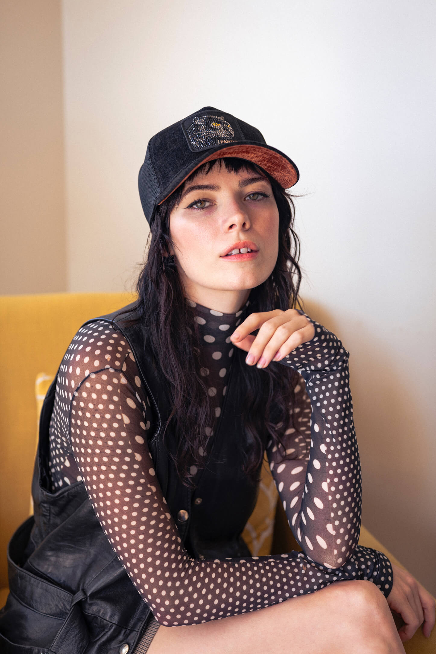 Woman wearing a black mesh polka dot top, black vest, and a Goorin black baseball cap sits on a yellow chair, looking at the camera with one hand raised near her face.