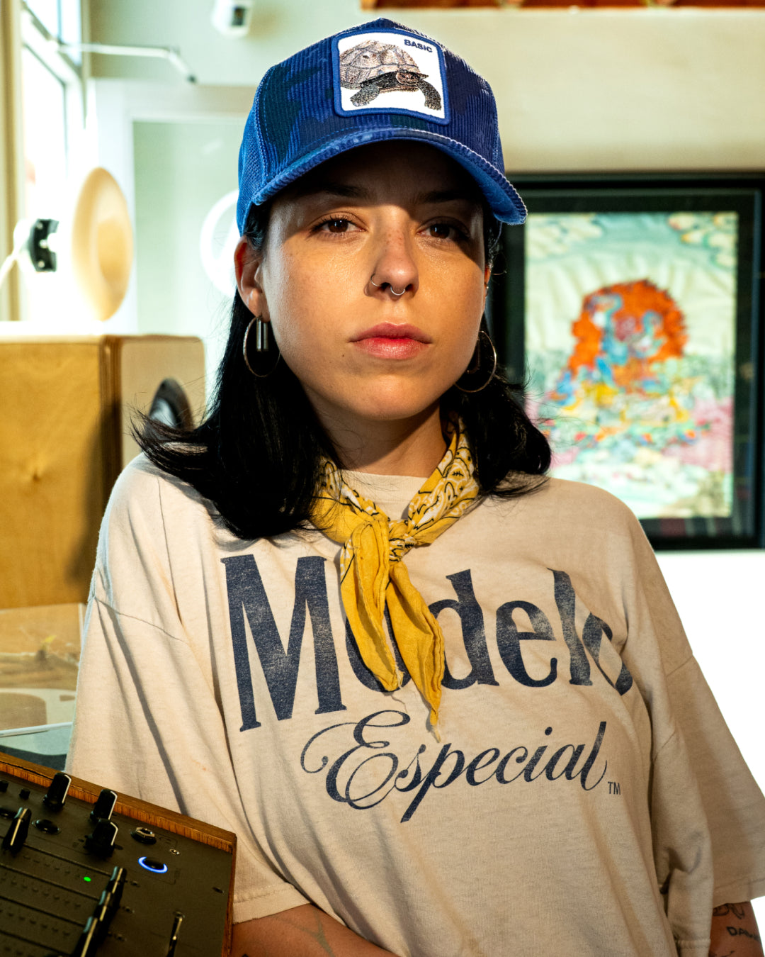A person wearing a bold hat design—a blue cap—hoop earrings, a yellow bandana, and a Modek Especial t-shirt stands indoors next to audio equipment.