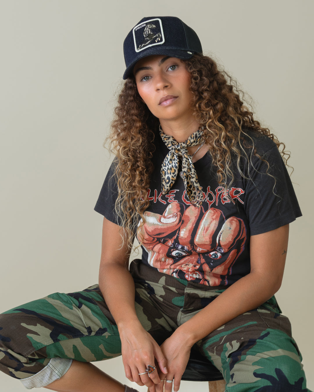 A woman with curly hair wears a black, American made cap, leopard print scarf, graphic T-shirt, and camouflage pants while sitting on a stool against a plain background.