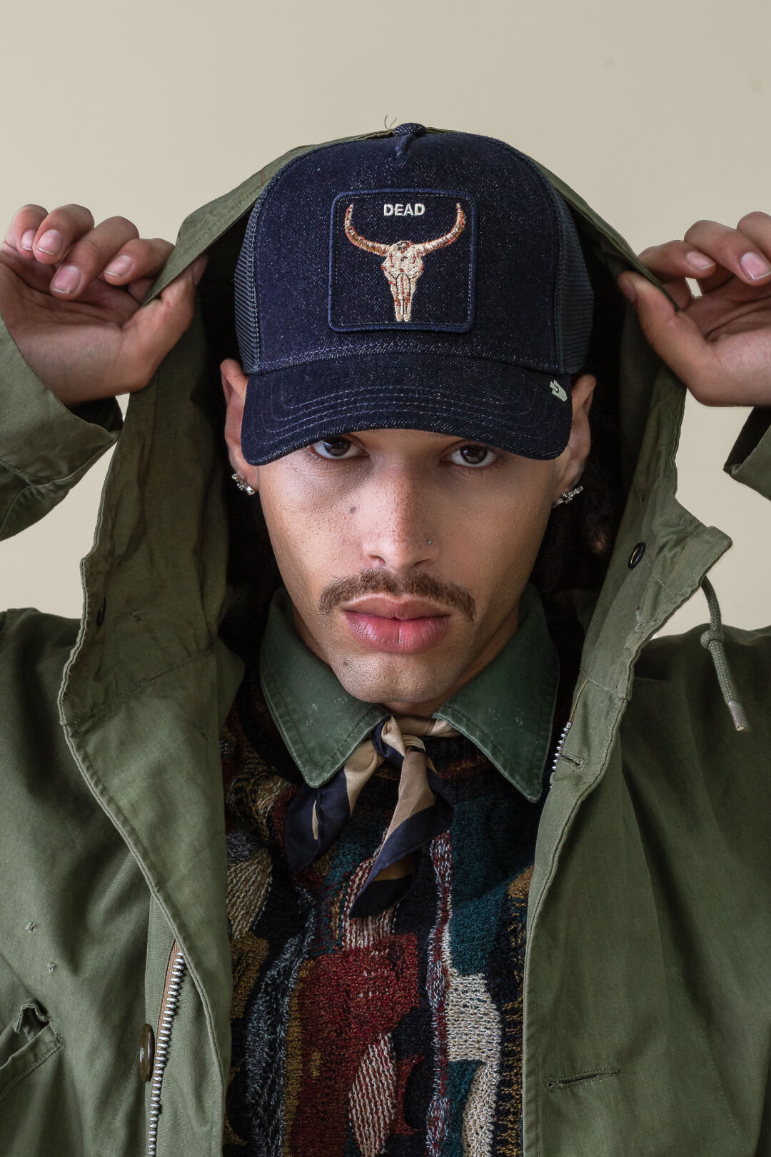 A person wearing a green jacket, patterned sweater, and a bold hat design—a black trucker hat with a bull skull and the word DEAD—holds the hood of their jacket.