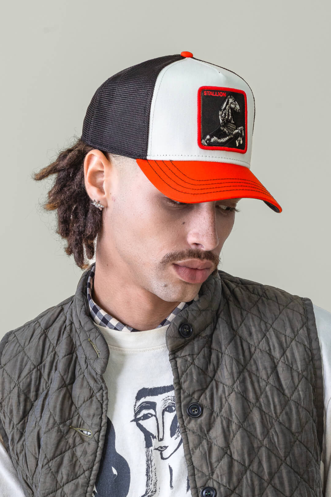 A person wearing a bold, American made red and white trucker hat with a Stallion patch, a quilted vest, and a shirt with a graphic face print, looking downward.