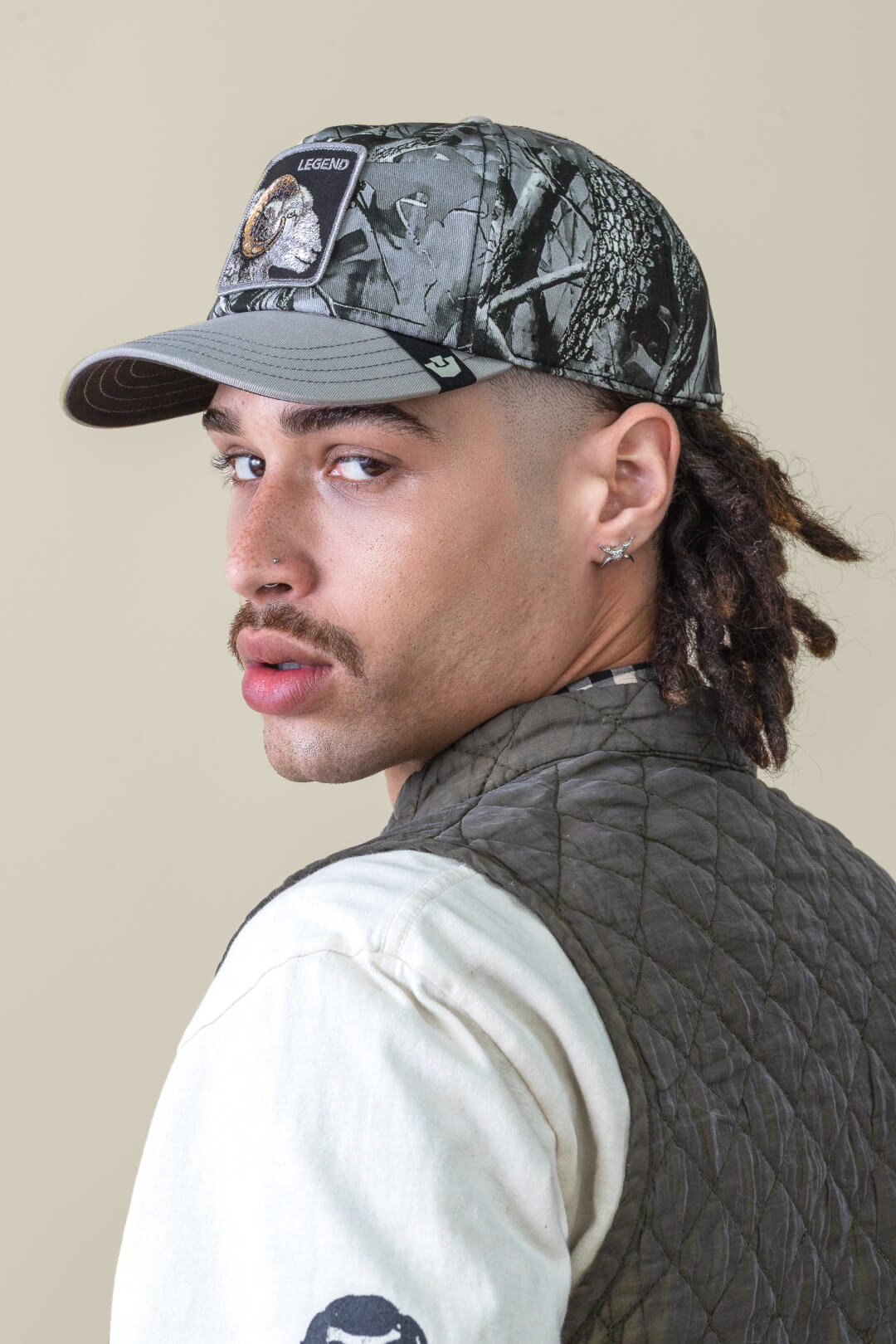 A person with a mustache and goatee wears a Goorin camo trucker hat, quilted vest, and cream shirt, looking over their shoulder at the camera against a plain background.