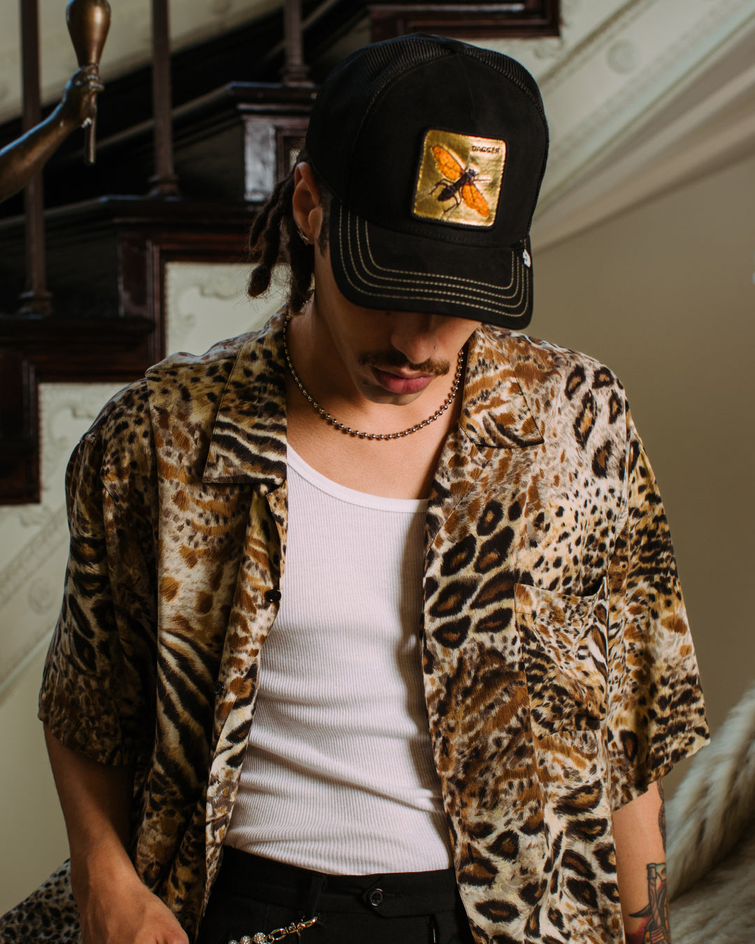 A person in a leopard print shirt, white tank top, and black cap with a butterfly patch—one of our durable hats—stands indoors near a staircase, looking downward.