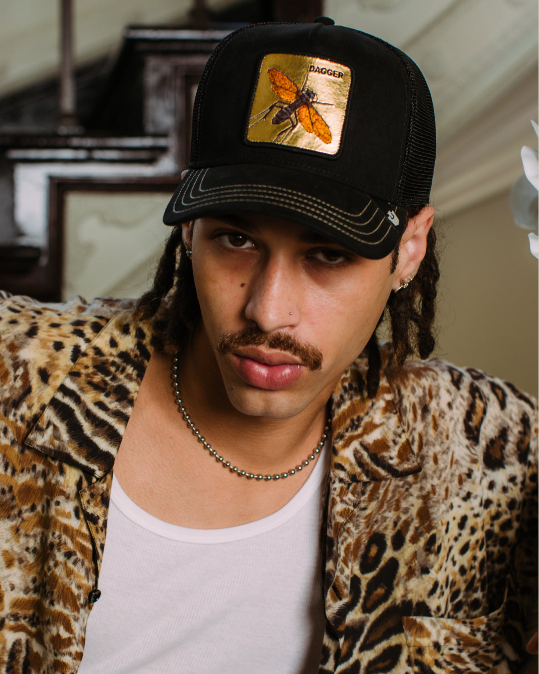 A man with a mustache wearing a Goorin black mesh hat with an insect patch, a leopard print shirt, a white tank top, and a beaded necklace sits indoors.