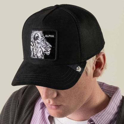 A person with light hair, dressed in a collared shirt and jacket, looks downward while wearing the Goorin Bros. “Black Out Lion” mesh stealth trucker hat, which features an embroidered lion patch labeled ALPHA.