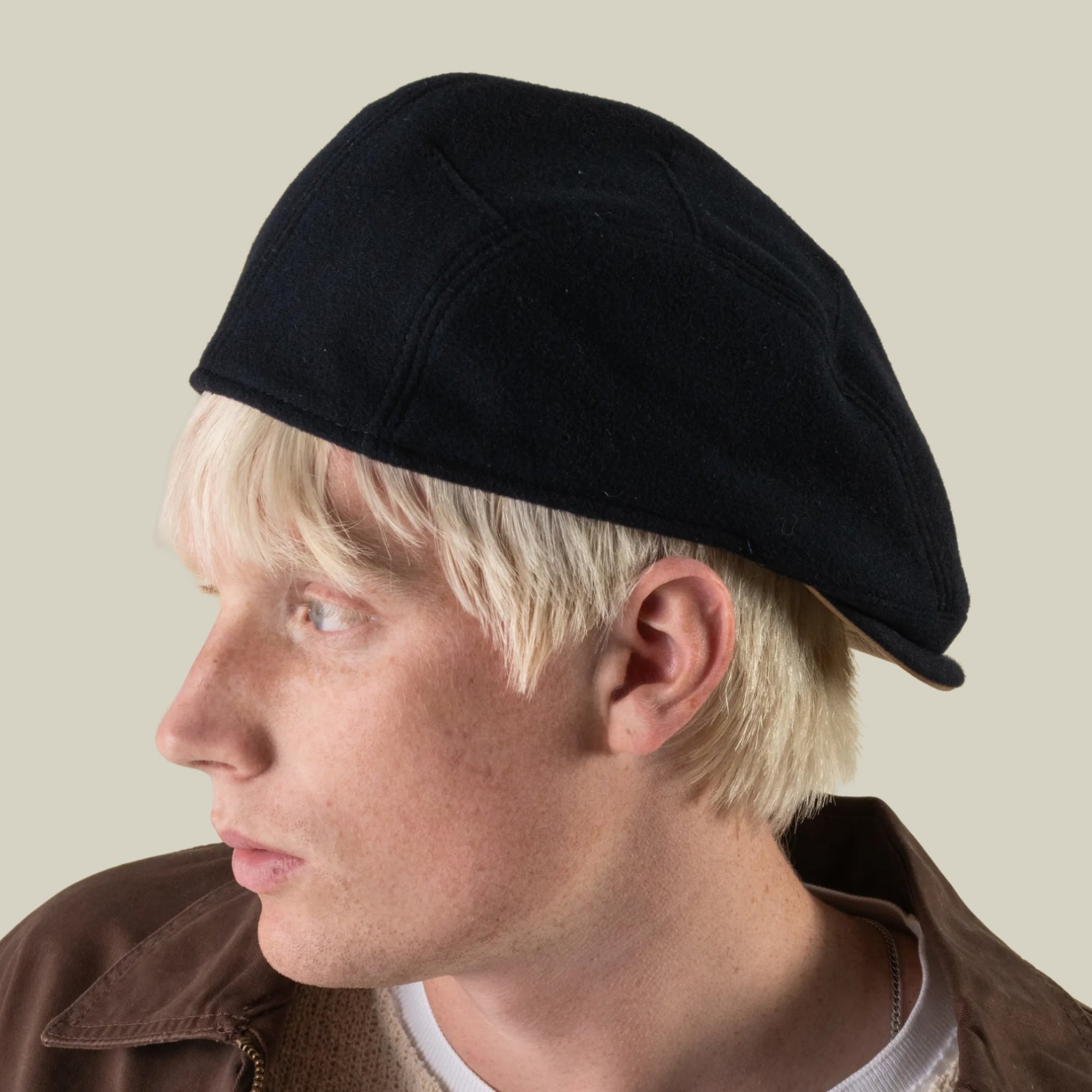A person with light blonde hair is shown in profile against a plain beige background, wearing the “Cornerstone” wool cap by Goorin Bros. and a brown jacket.