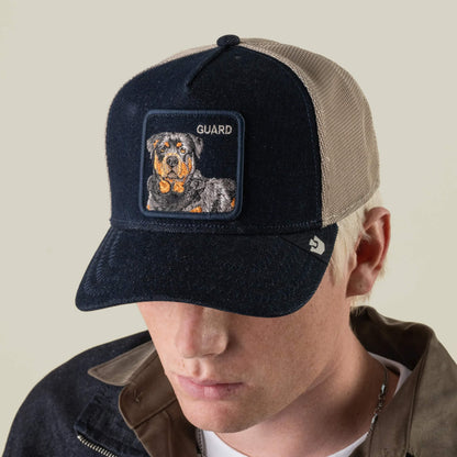 A person wearing the Goorin Bros. “Denim Guard” two-tone denim trucker hat with an embroidered Rottweiler patch and the word GUARD on the front, looking down.