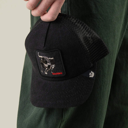A person holds the Goorin Bros. Denim Raging breathable mesh trucker hat, featuring a snake patch with the word “RAGING” in red, next to dark green pants.