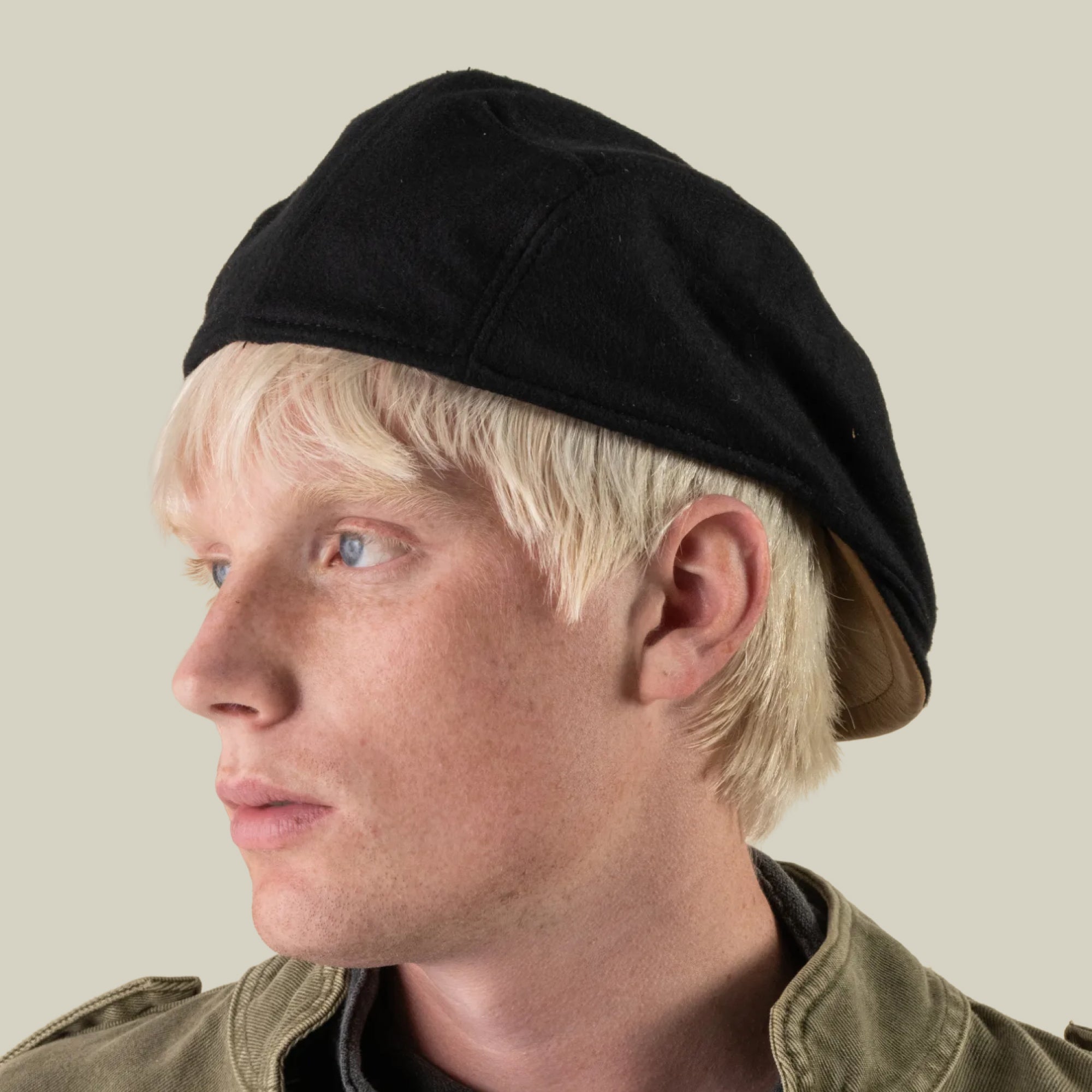 A person with light blonde hair and fair skin looks to the left, wearing the Cornerstone black wool cap by Goorin Bros., handmade in New Jersey, along with a green jacket against a plain, light background.