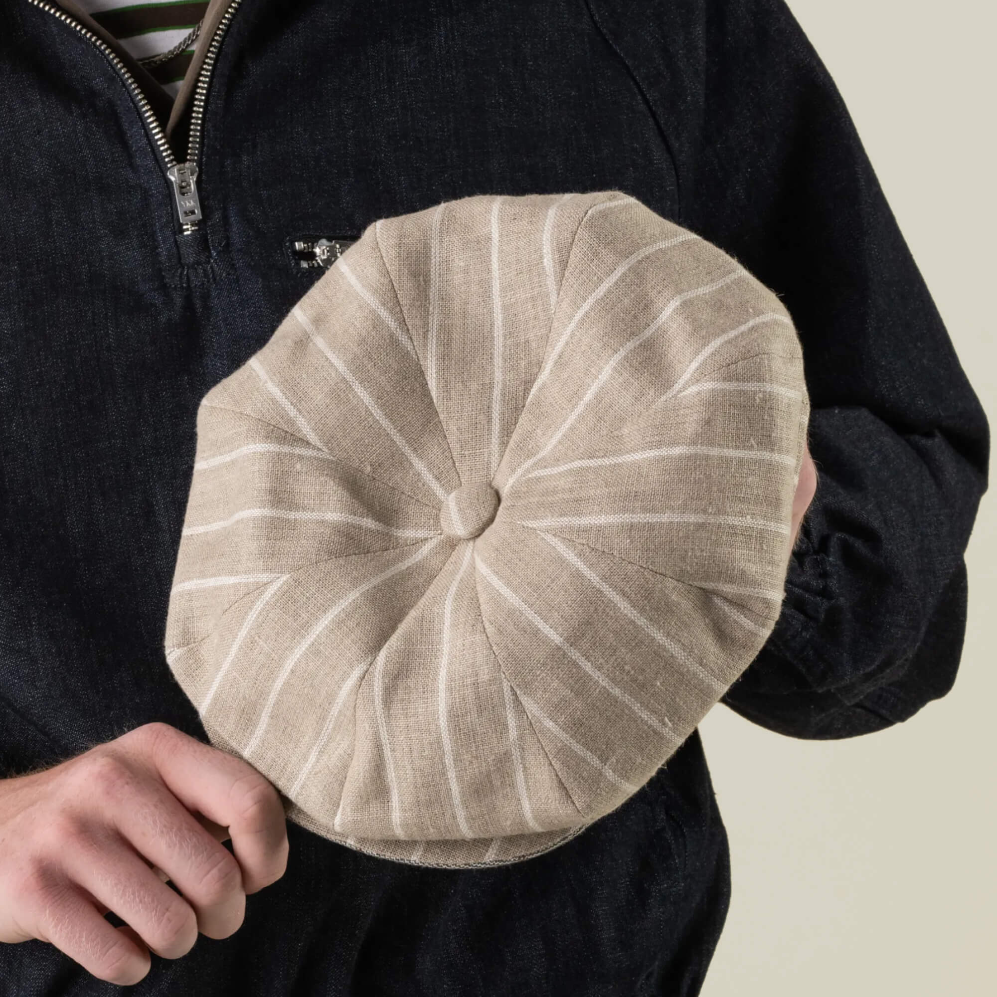 A person holds the Rincon Park cap by Goorin Bros.—a beige, round, pinstriped hat with a button top, hand cut and sewn in a classic Gatsby silhouette—while wearing a dark zip-up jacket.