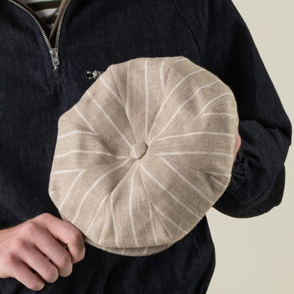 A person holds the Rincon Park cap by Goorin Bros.—a beige, round, pinstriped hat with a button top, hand cut and sewn in a classic Gatsby silhouette—while wearing a dark zip-up jacket.