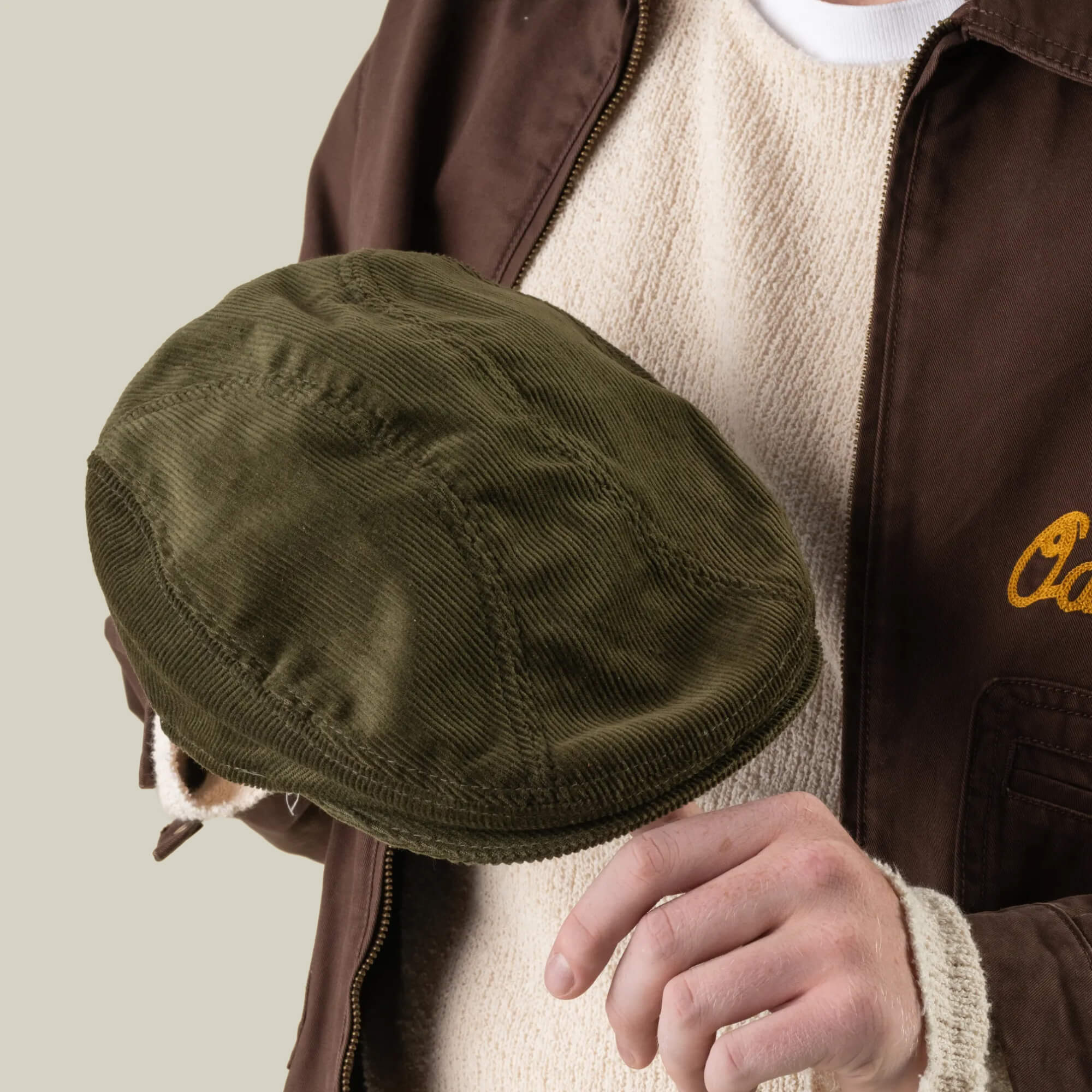 A person wearing a beige sweater and brown jacket holds the Maxwell Park olive green corduroy hat by Goorin Bros. in one hand.