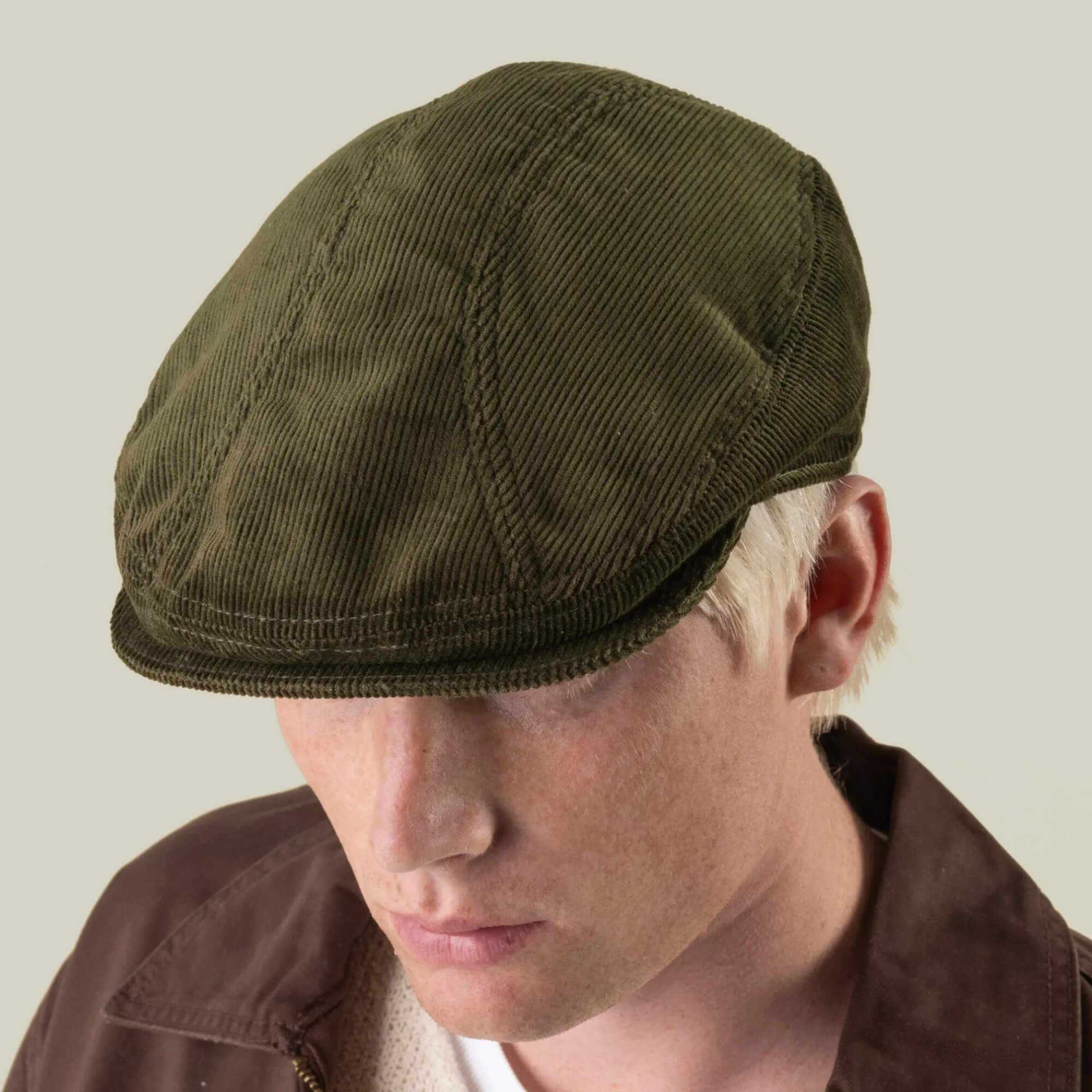A person wearing the Maxwell Park by Goorin Bros.—a durable, expertly hand-cut olive green corduroy flat cap made in the USA—paired with a brown jacket, facing slightly downward against a plain background.