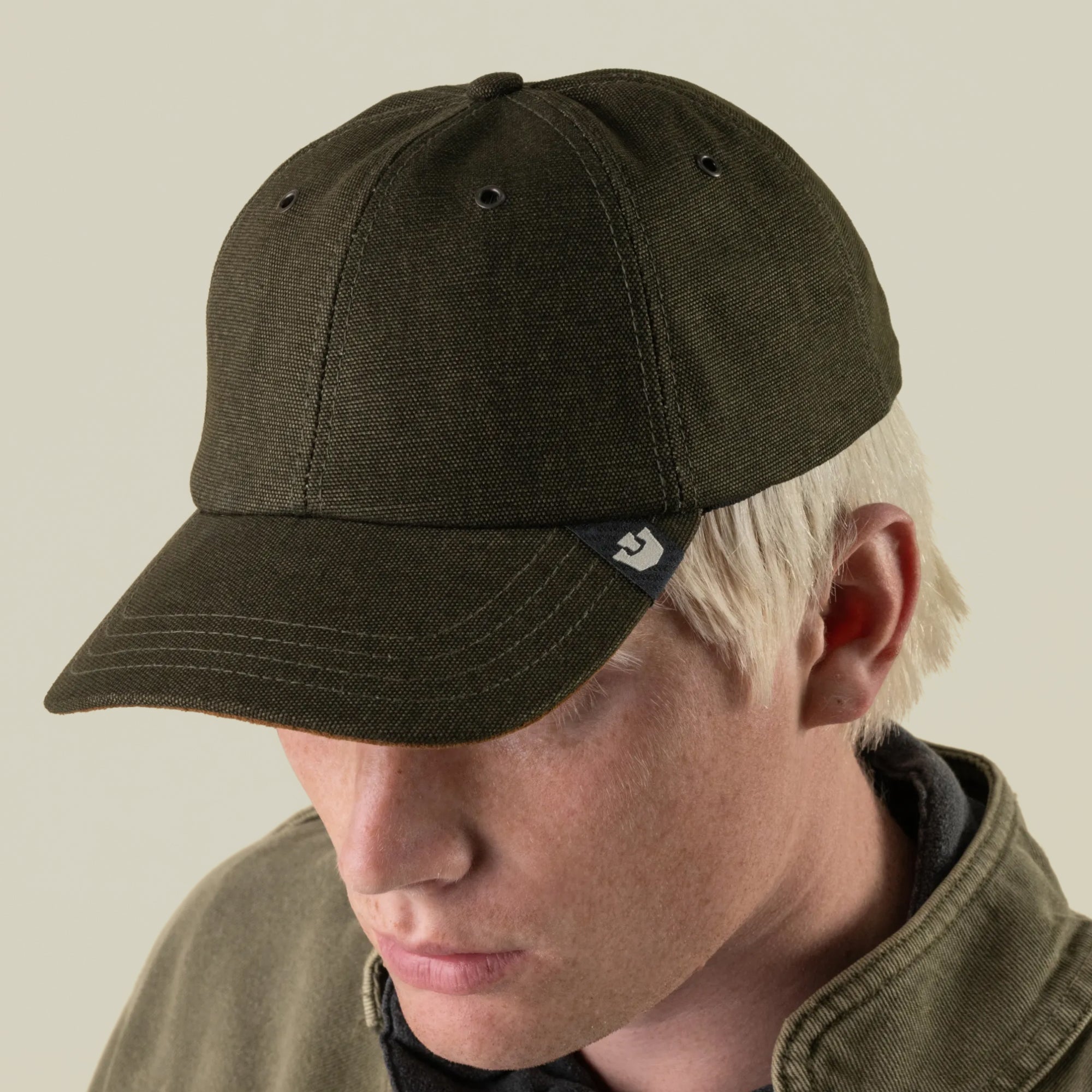 Someone with light hair wears the Goorin Bros. Mineral Road mixed-material dark olive baseball cap and a matching jacket, looking down against a plain background.