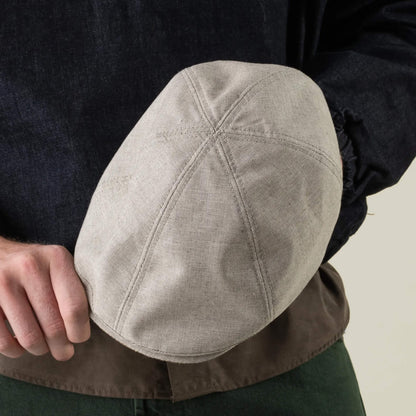 A person holds the Goorin Bros. Mile Rock— a light gray, hand cut and sewn 6 Panel Ivy flat cap with visible stitching, made in the USA— while wearing a dark top and green pants.